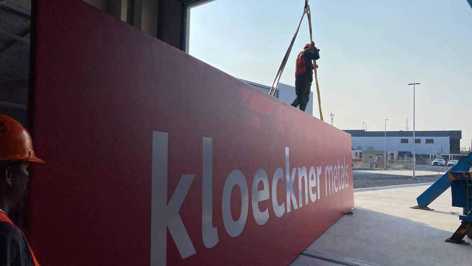 Construction workers installing a large red sign with white lettering that reads "Kloekner markets" outside a building at a commercial construction site.