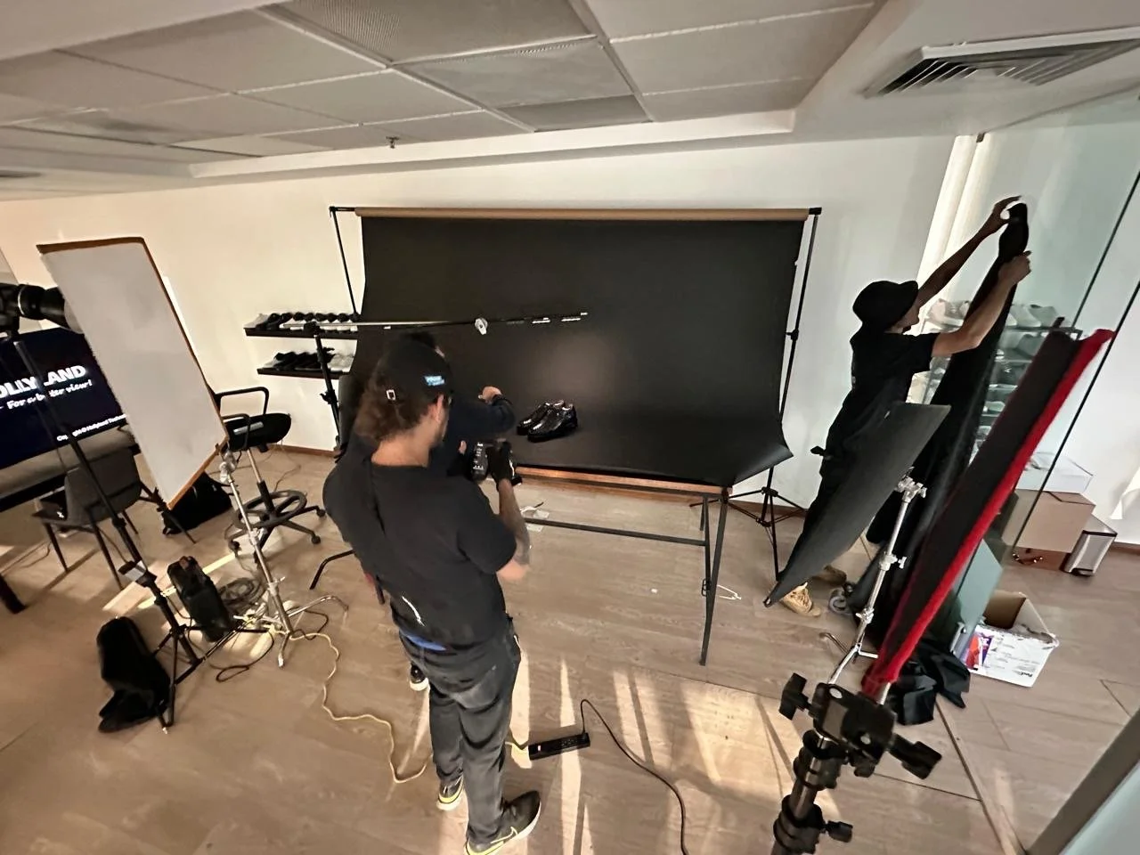 Photography studio with backdrop and lighting equipment where two people are setting up for a shoot; one person adjusting items on a black backdrop, another organizing lights and gear.