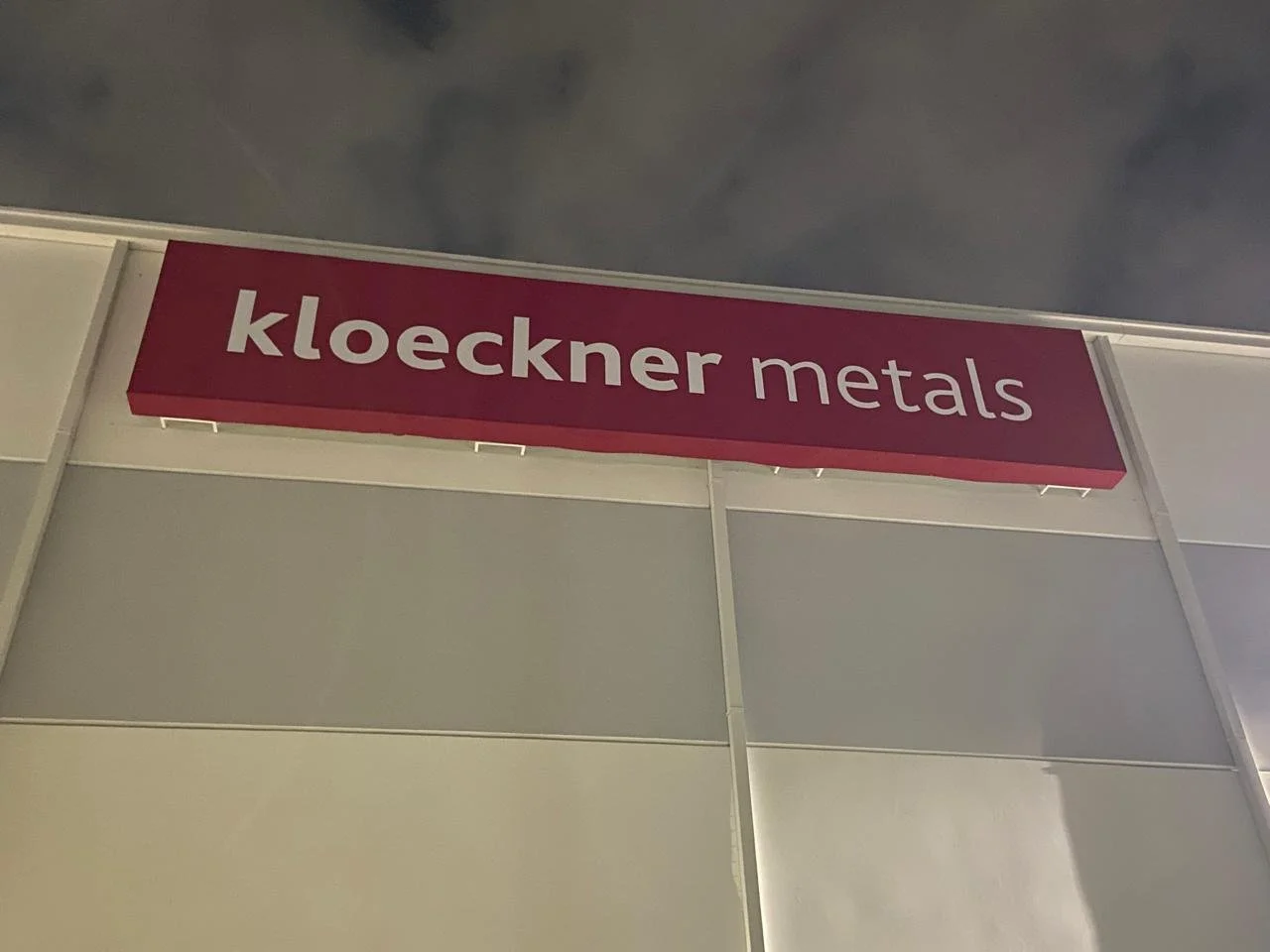 Signboard that reads 'Kloeckner metals' in white letters on a dark pink background, mounted on an exhibition booth with a grey wall and a cloudy sky background.