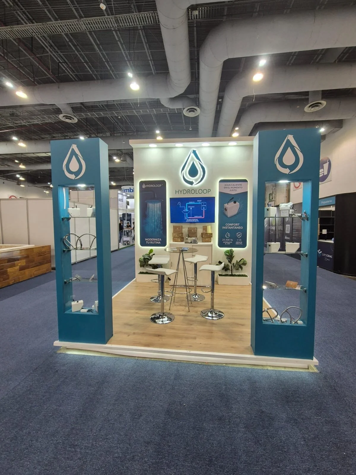 Trade show booth for Hydrolloop, featuring a blue and white color scheme, with display shelves showcasing water-related products, a central area with high chairs and a small table, and posters about water heating and instant comfort.