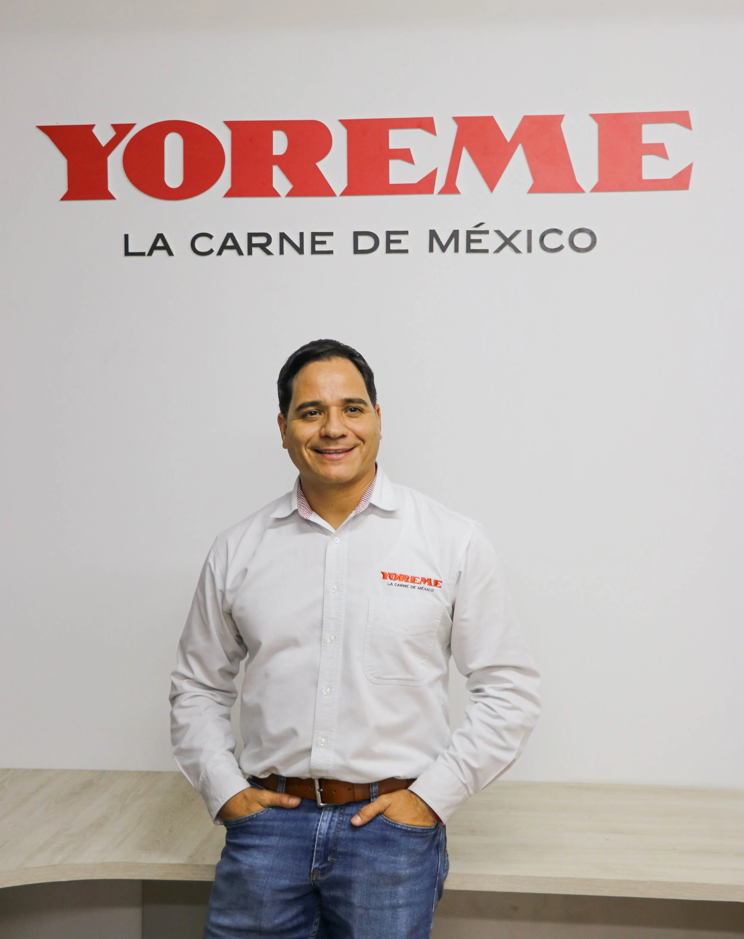 Man standing in front of a wall with the text 'YOREME LA CARNE DE MÉXICO', wearing a white shirt with the Yoreme logo, smiling, with hands in pockets.