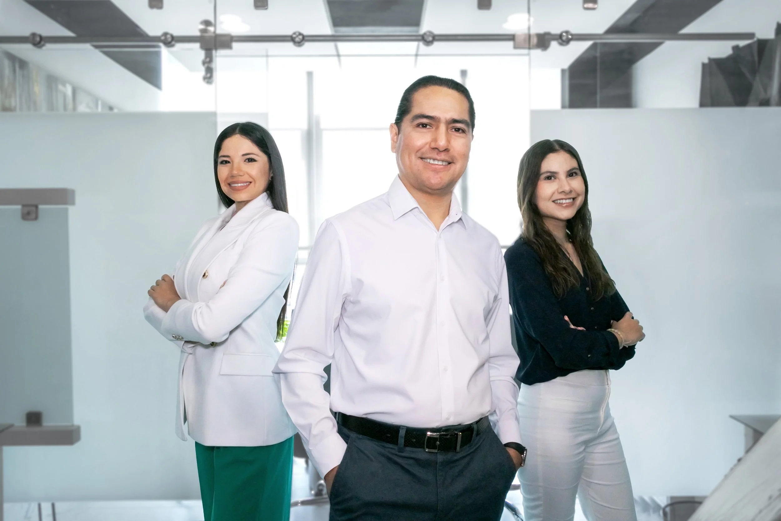 Three professionals standing in an office, with two women and one man, smiling and dressed in business casual attire.