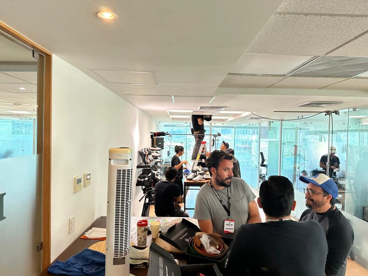People working inside a glass-walled office space, some seated and others standing, with cameras and equipment, suggesting a behind-the-scenes setup or filming session.