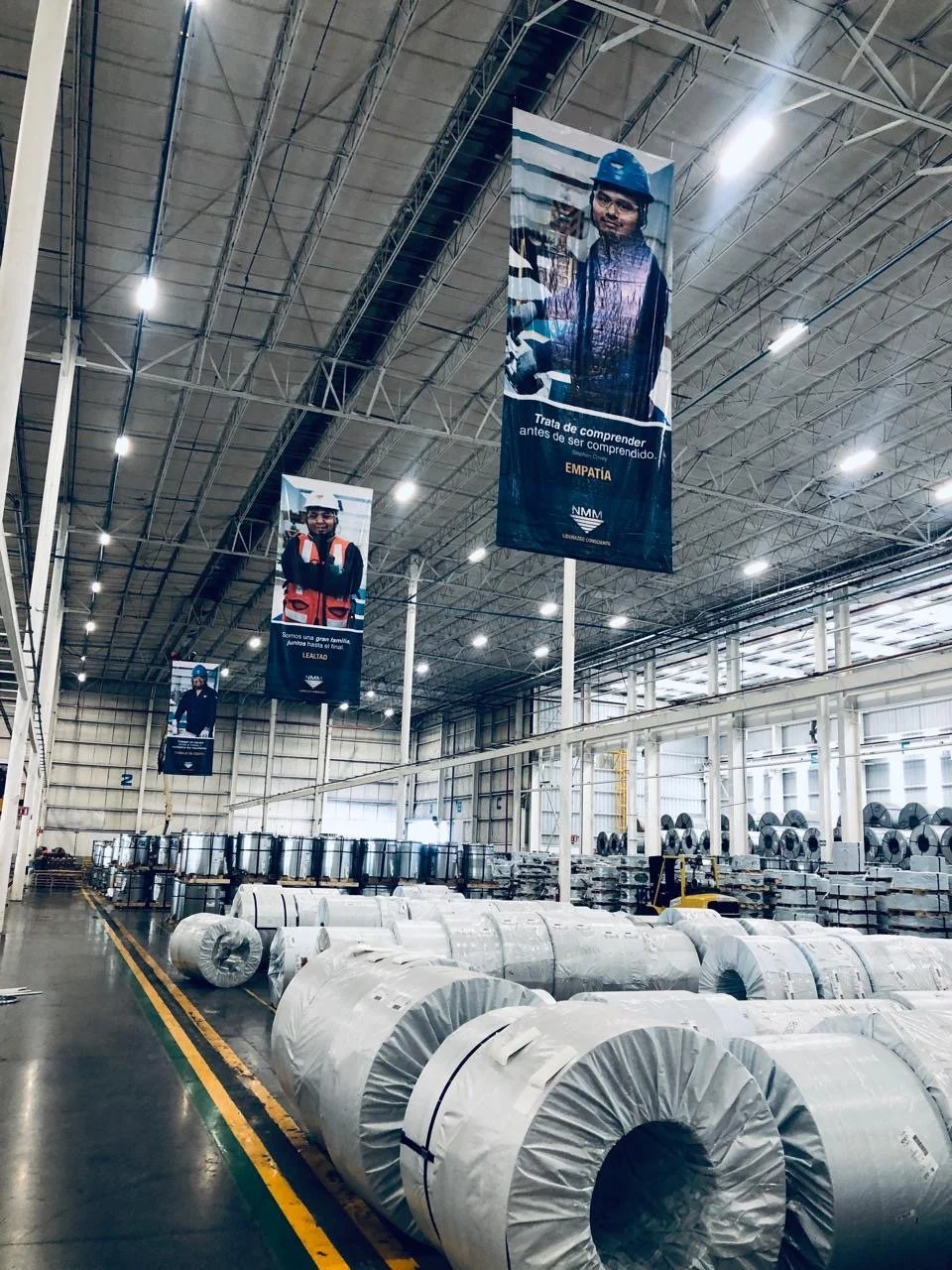 Inside a large industrial warehouse with rolls of steel or metal stacked on the floor. There are banners hanging from the ceiling with images of workers and messages promoting understanding and empathy.
