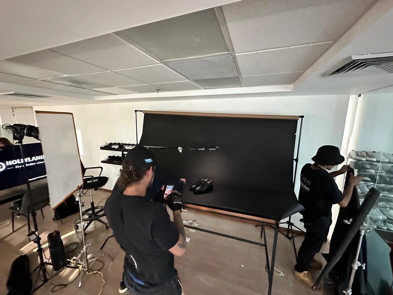 Indoor photo of a photography studio setup with a black backdrop, lights, and equipment. Two people are working; one is taking a photo, and the other is adjusting items nearby.