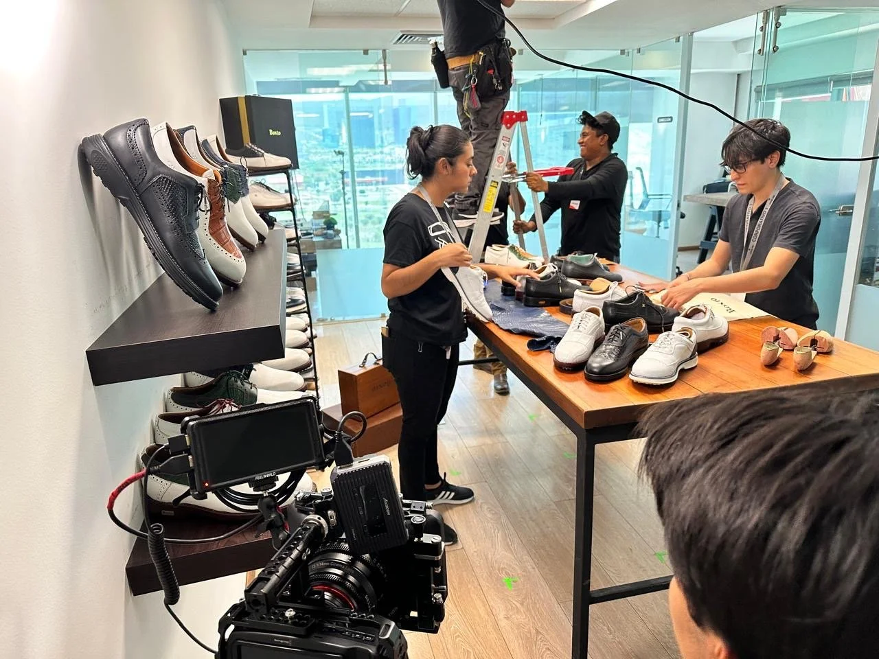 People filming a shoe commercial in a modern indoor space with glass walls, displaying various sneakers on a table and shelves, filming equipment, and a person on a ladder in the background.