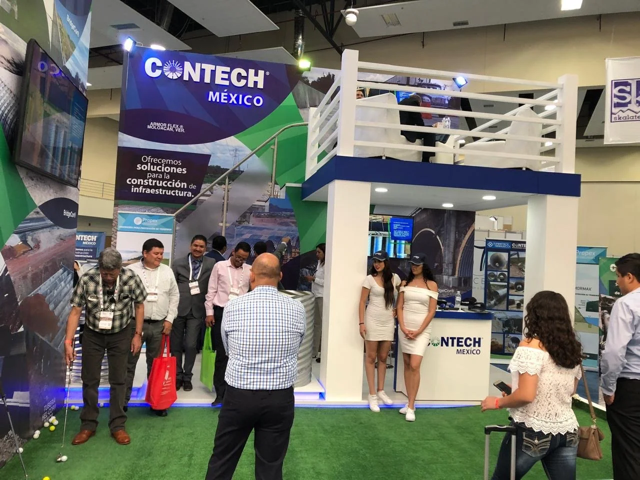 Trade show booth for CONTECH MÉXICO featuring a small elevated platform, branding banners, and a golf putting green with several people participating in a golf activity, surrounded by visitors and promotional staff.