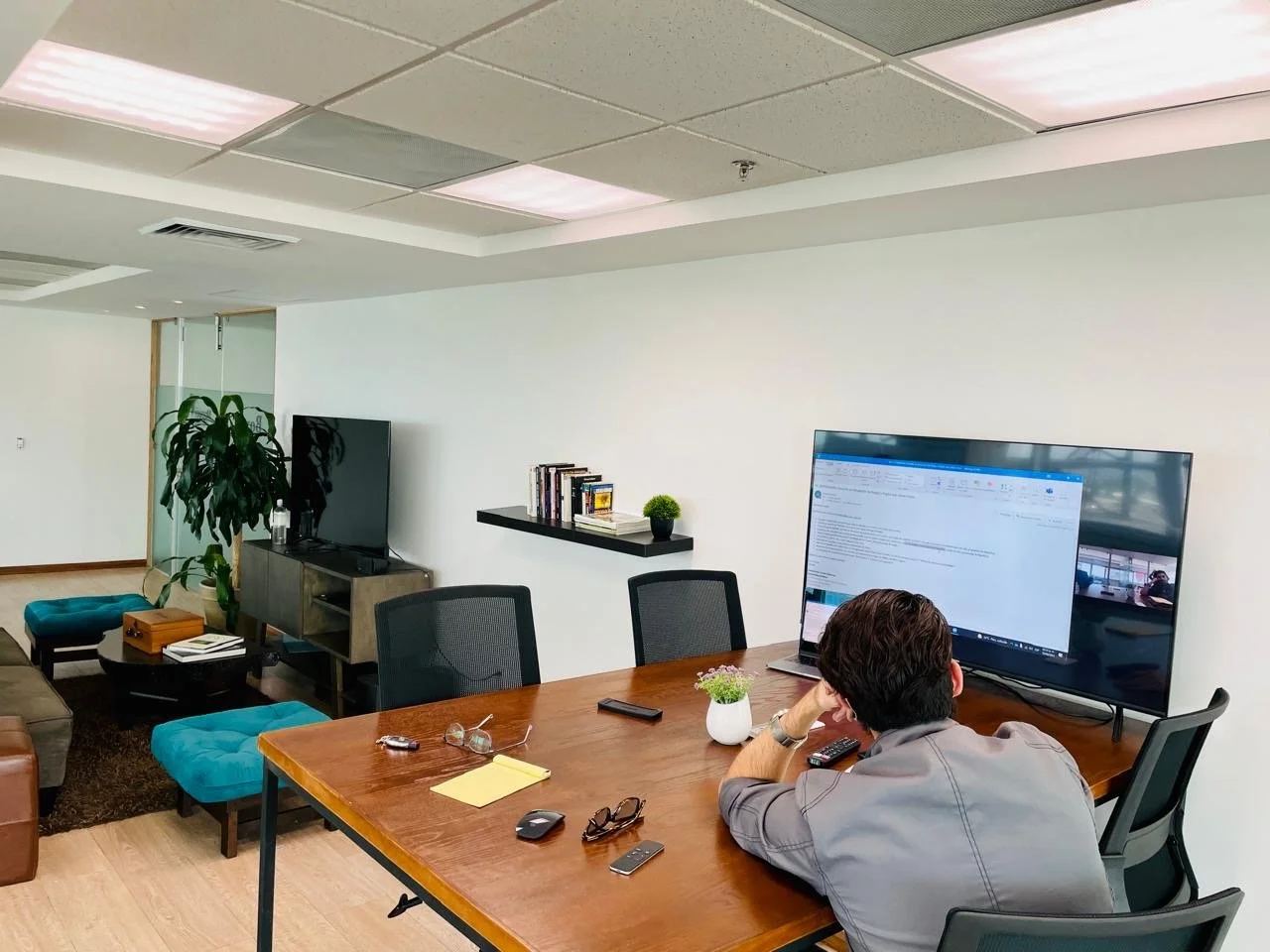 A person working at a wooden desk with computer monitors, remote controls, eyeglasses, and a notepad. The room has a large TV, bookshelf, potted plant, and sitting area with chairs and a sofa.
