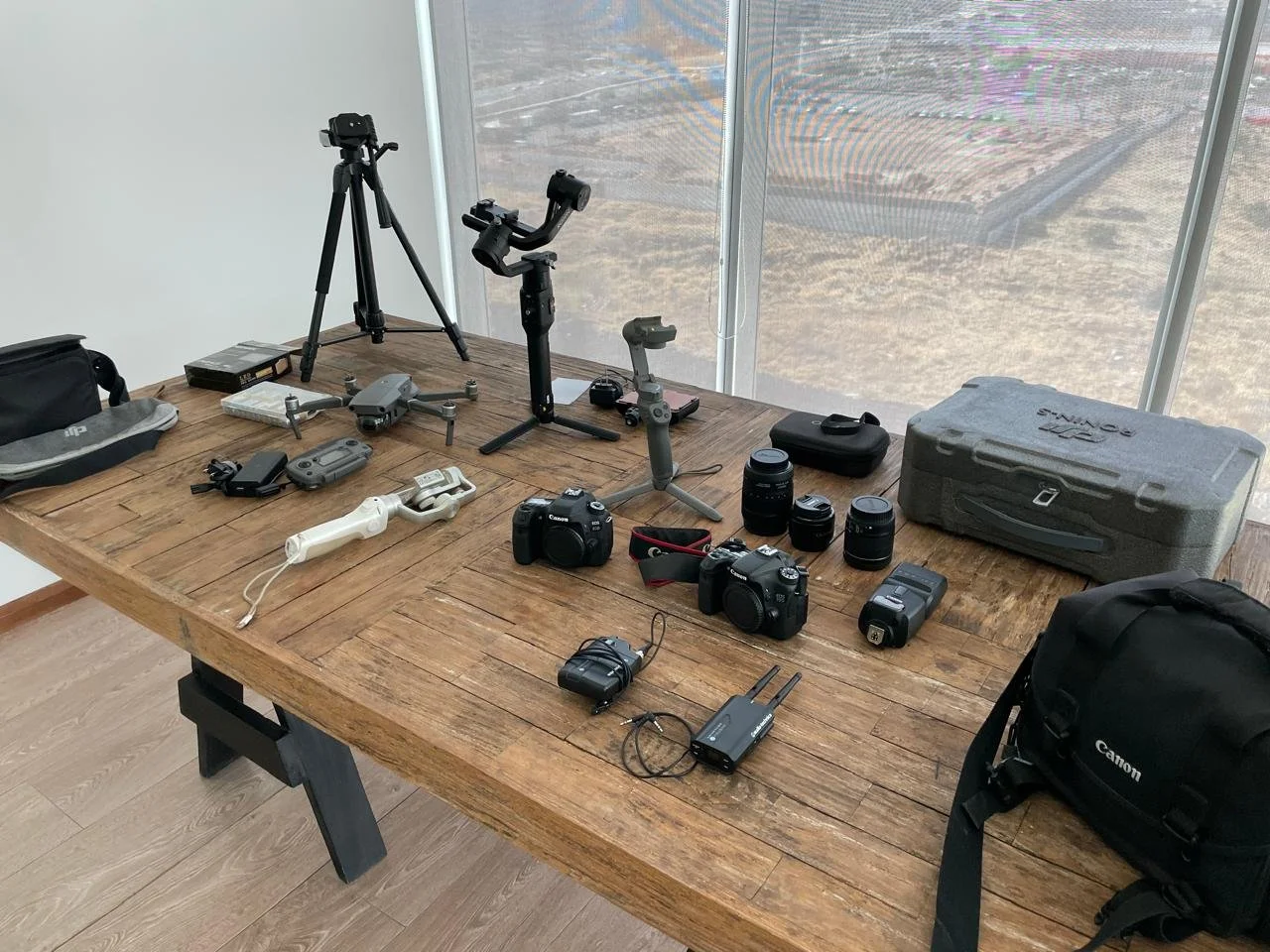 Table with various camera equipment including tripods, cameras, lenses, batteries, and accessories in front of a large window with a cityscape view.