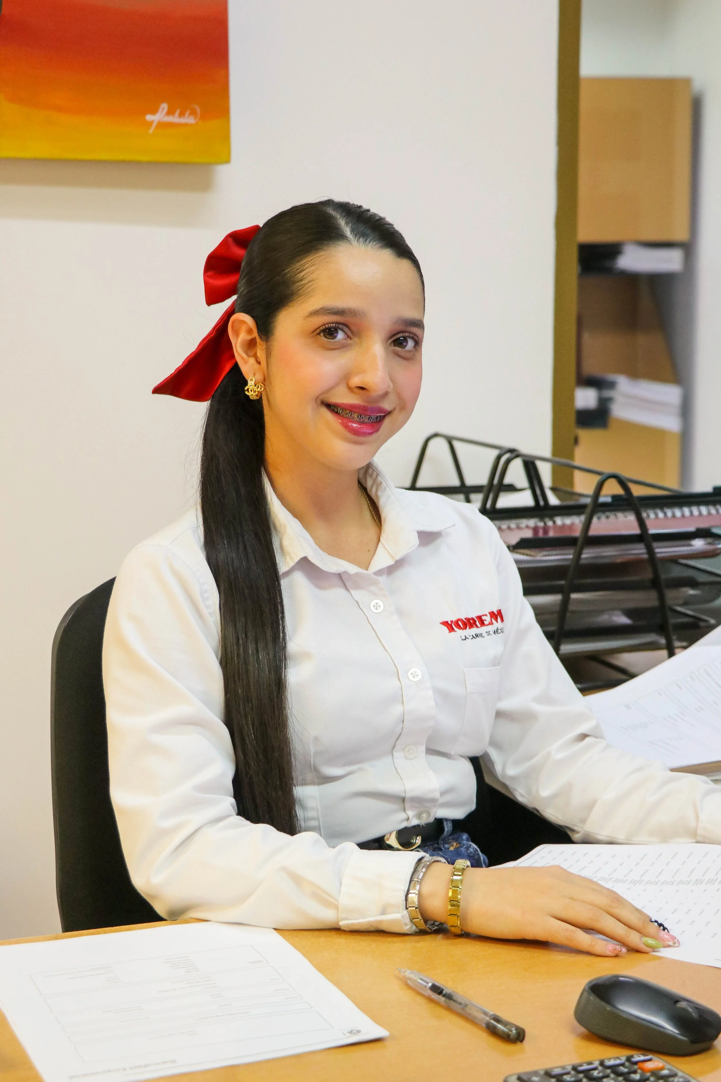 A young woman with long dark hair, sitting at a desk, wearing a white shirt with red embroidery, with a red ribbon in her hair, smiling, surrounded by papers, pens, and office supplies in an office setting.