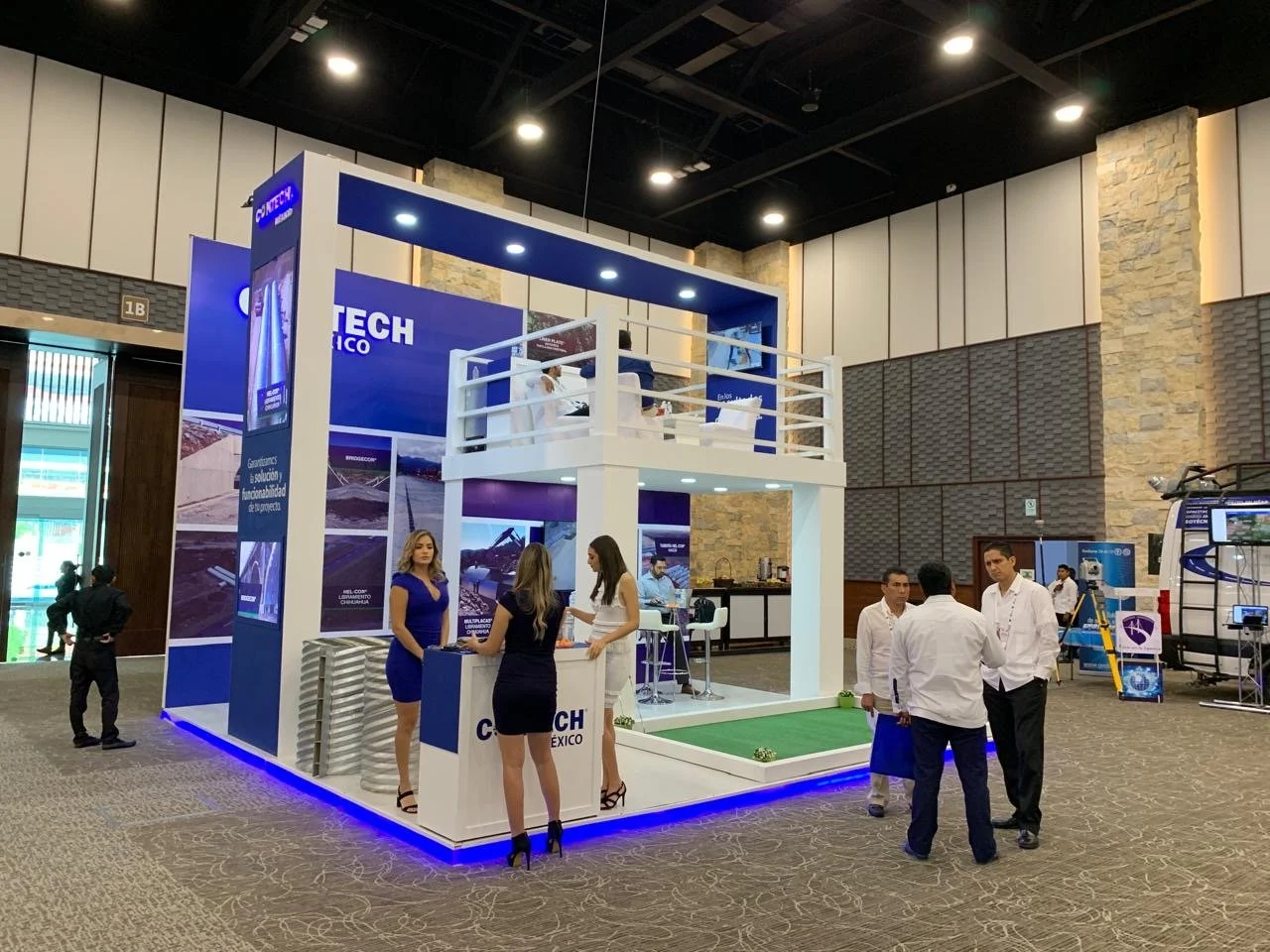 Trade show booth with blue and white design, featuring the text 'CONTECH Mexico'; includes a second-floor balcony, seating area, and a small artificial grass area with a mini-golf putting green; several people are talking and walking around.