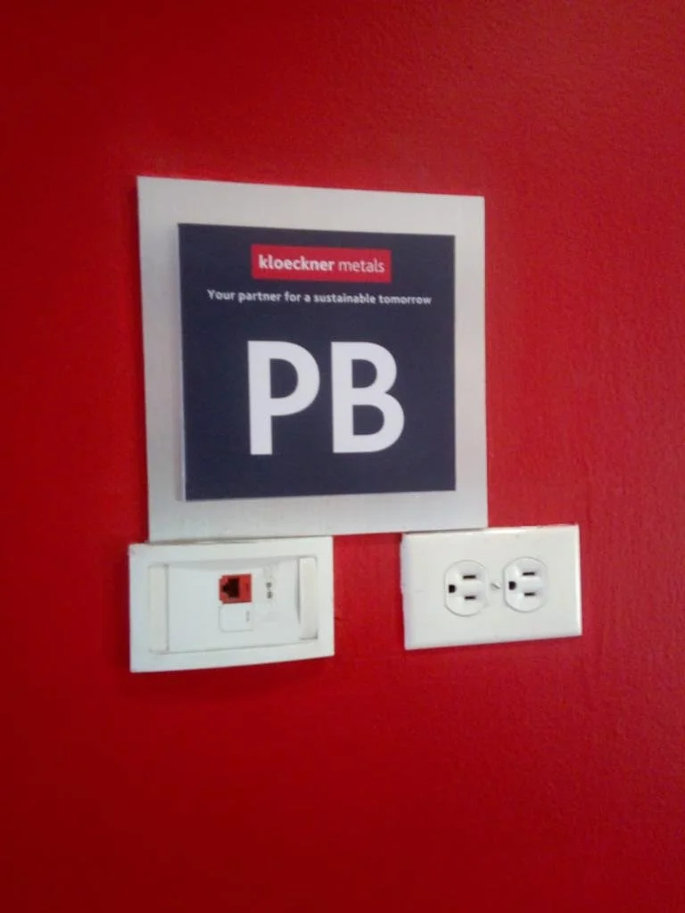 A poster on a red wall with the text 'kloeckner metals' and 'Your partner for a sustainable tomorrow' above the symbol for lead (Pb). Below the poster are a white electrical outlet and a white switch plate.
