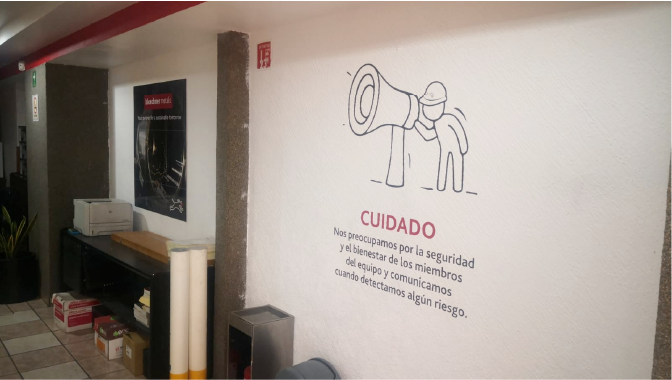 Wall with a black and white illustration of a person with a megaphone, and a red 'CUIDADO' warning sign in Spanish, in an indoor space with a printer, boxes, and a trash can.