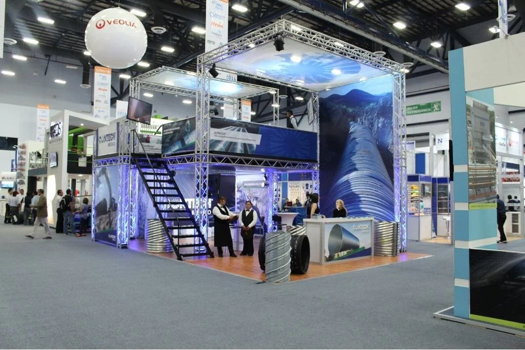 Trade show booth with metallic framework, large water pipeline images on walls, and staff members standing and talking at various tables.