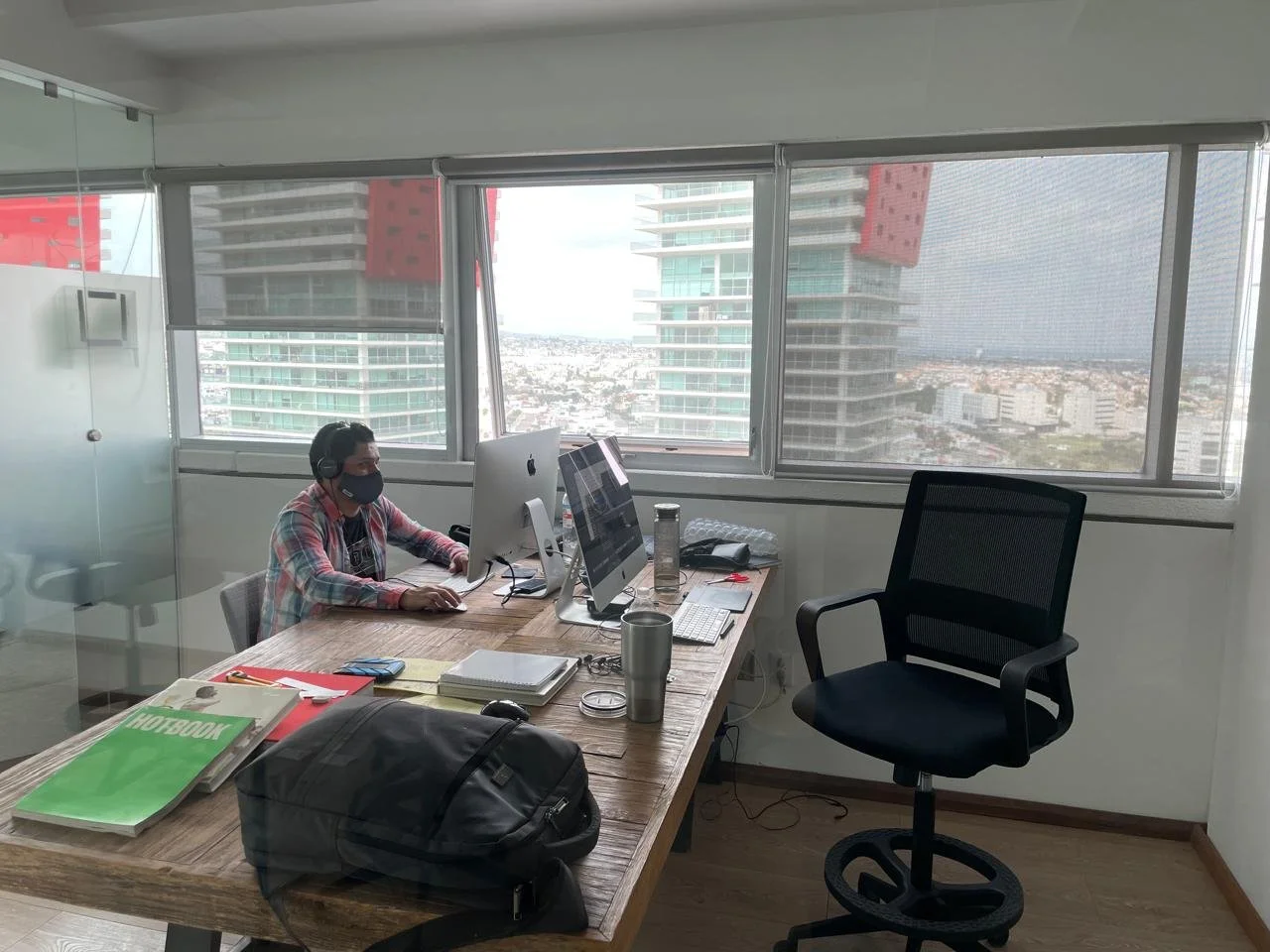 An office with a man wearing a face mask and headphones working on a computer, cluttered desk with notebooks, water bottles, and a backpack, large windows overlooking a city with tall buildings.