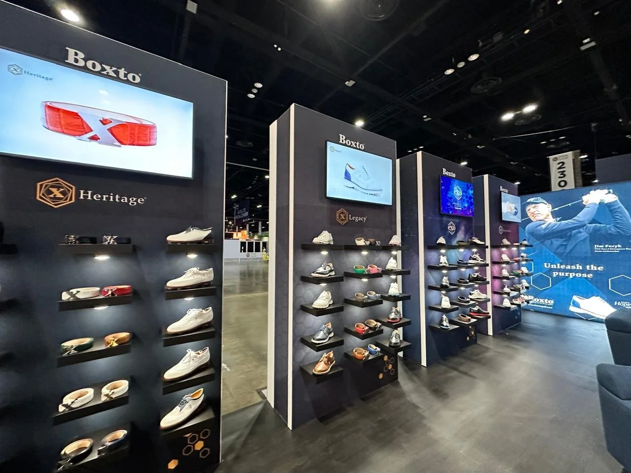 Exhibit of Atlanta's Boxto brand shoes at a trade show, including various athletic shoes and sneakers displayed on black shelves, with digital screens and promotional signage in the background.