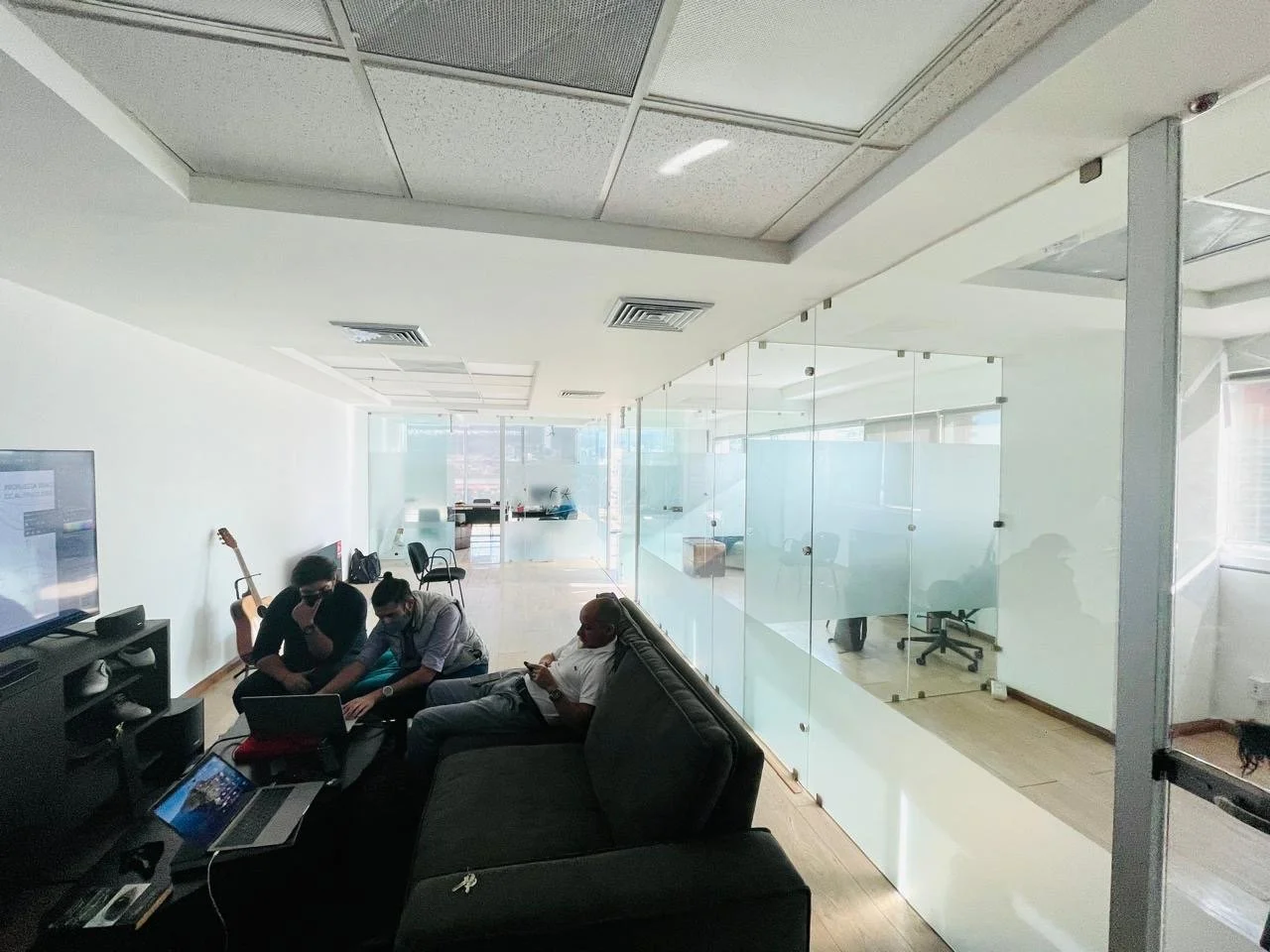 An office lounge area with three people sitting on a black sofa, two using laptops and one looking at their phone. There is a TV on a stand and two guitars leaning against the wall. Behind the glass partition, there is a workspace with chairs and des