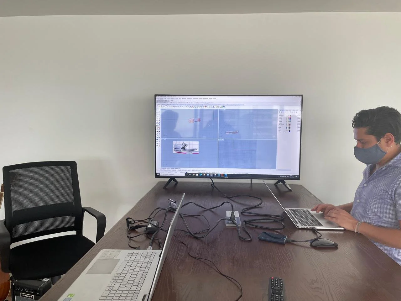 A person wearing a face mask working on a laptop at a desk with a large monitor displaying a 3D modeling program. An empty chair is nearby, and office supplies are on the desk.
