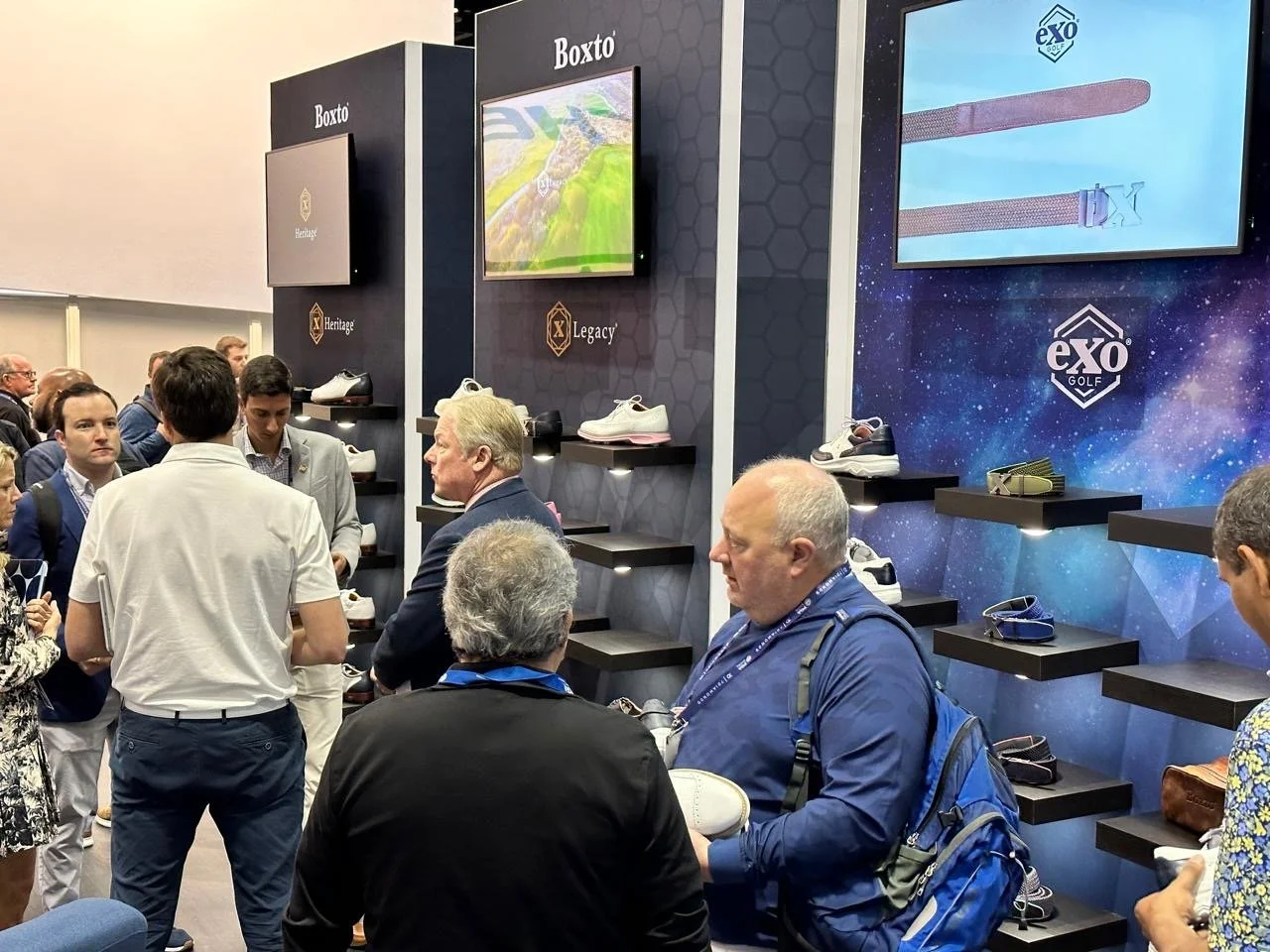 People gathered at a booth displaying golf shoes and accessories, with screens and branding for Exo Golf and legacy golf shoes.