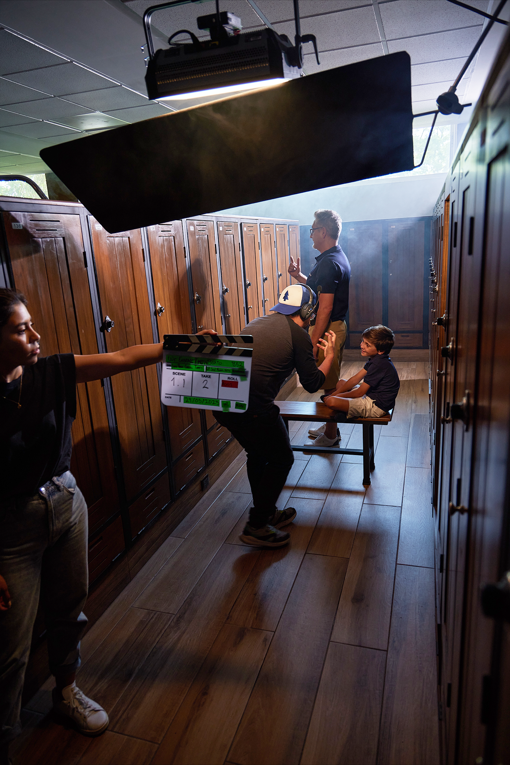 A scene on a film or video set showing a person sitting on a bench in a locker room, a director giving instructions, a crew member with a clapperboard, and a woman with a mic in front of the scene. The set is lit by professional lighting equipment ab