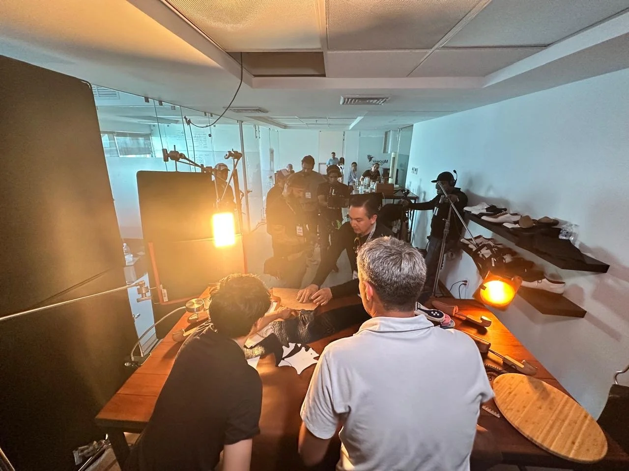 Filming crew setting up lighting and equipment on a set, with people around a table and some behind the camera, in a room with glass walls and ceiling lights.
