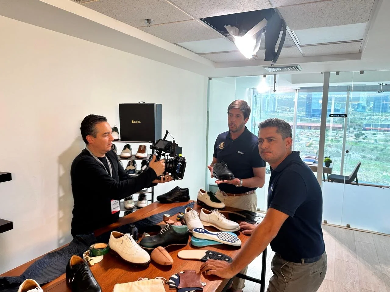 Two men discussing shoes at a shoe display table while a man films them with a camera in a modern office setting with a view of city buildings outside.