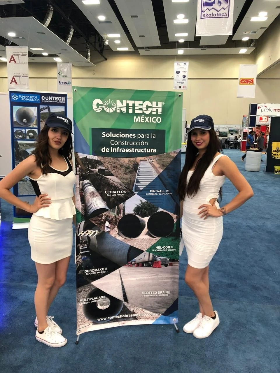 Two women standing on either side of a green and black CONTECH Mexico promotional banner at an indoor exhibition. Both women are wearing white dresses and navy blue caps with the CONTECH logo, smiling and posing for the photo.