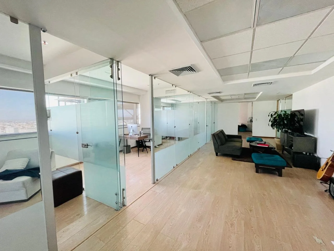 Modern office space with glass partitions, a seating area with a TV, couch, and chairs, and a small workspace with a computer.