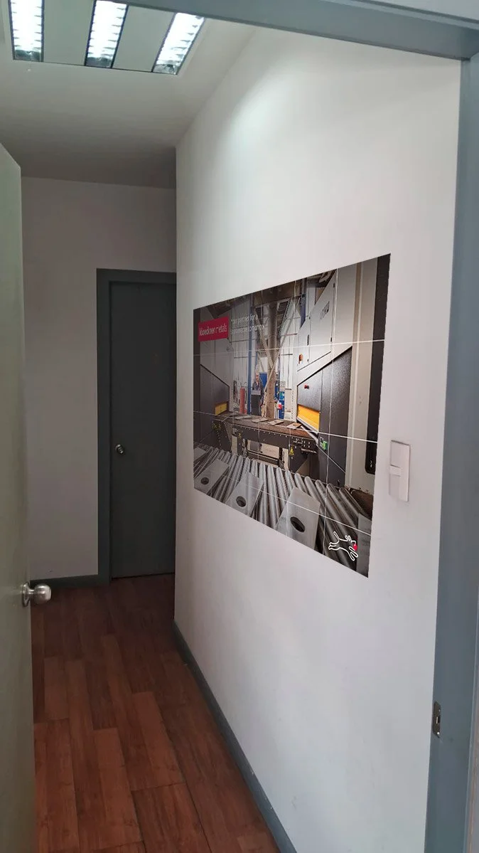 Interior of an office with a hallway, white walls, gray door, wooden floor, and a large wall-mounted digital display showing machinery in a manufacturing setting.