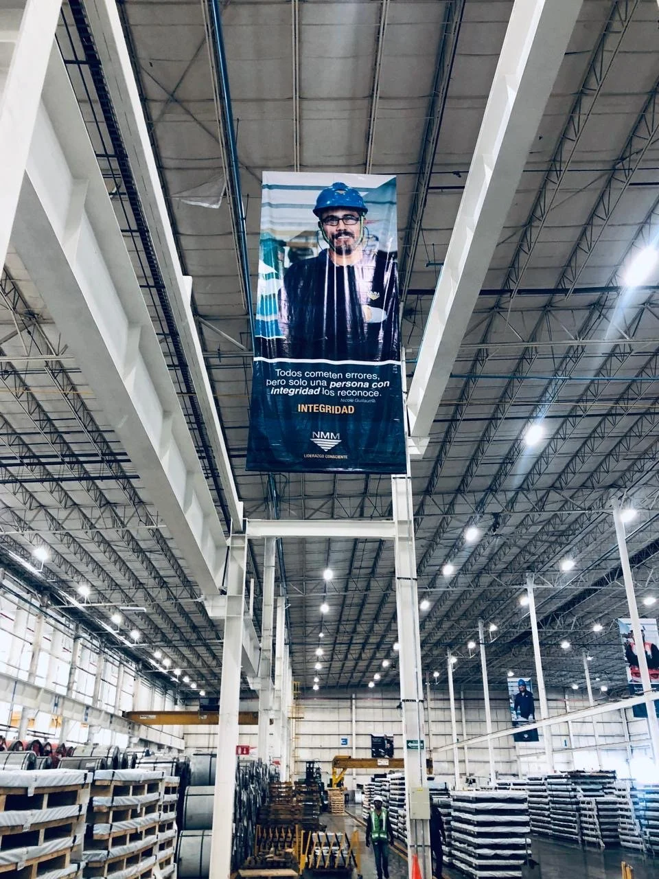 Large industrial warehouse with high ceilings and bright lighting, displaying vertical banners with a man wearing glasses, a helmet, and a safety vest. Shelves and equipment are visible in the background.