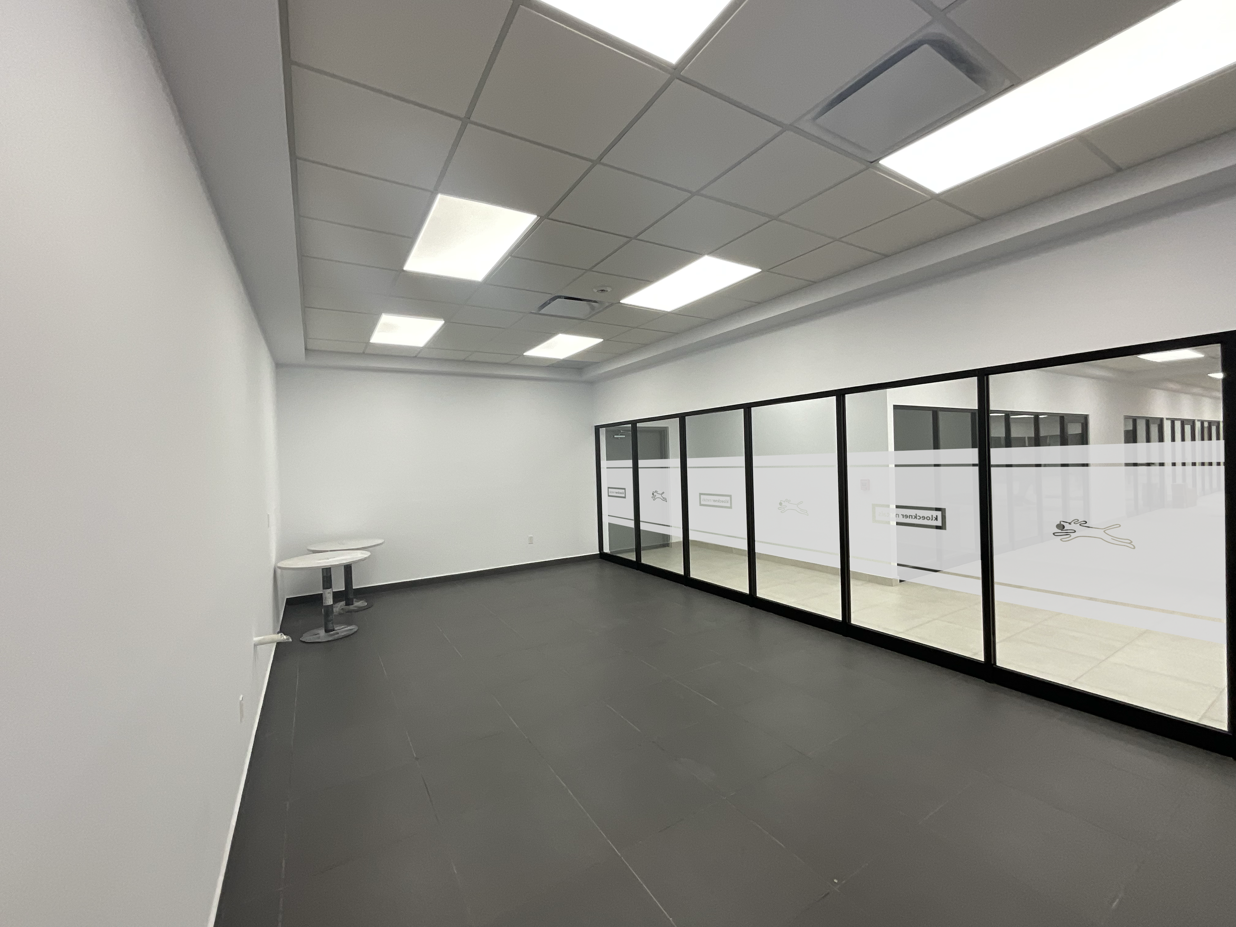 Empty office room with black tiled floor, white walls, and ceiling with fluorescent lights. There are two small round tables in the corner and a glass partition with frosted logos separating the space from another room.