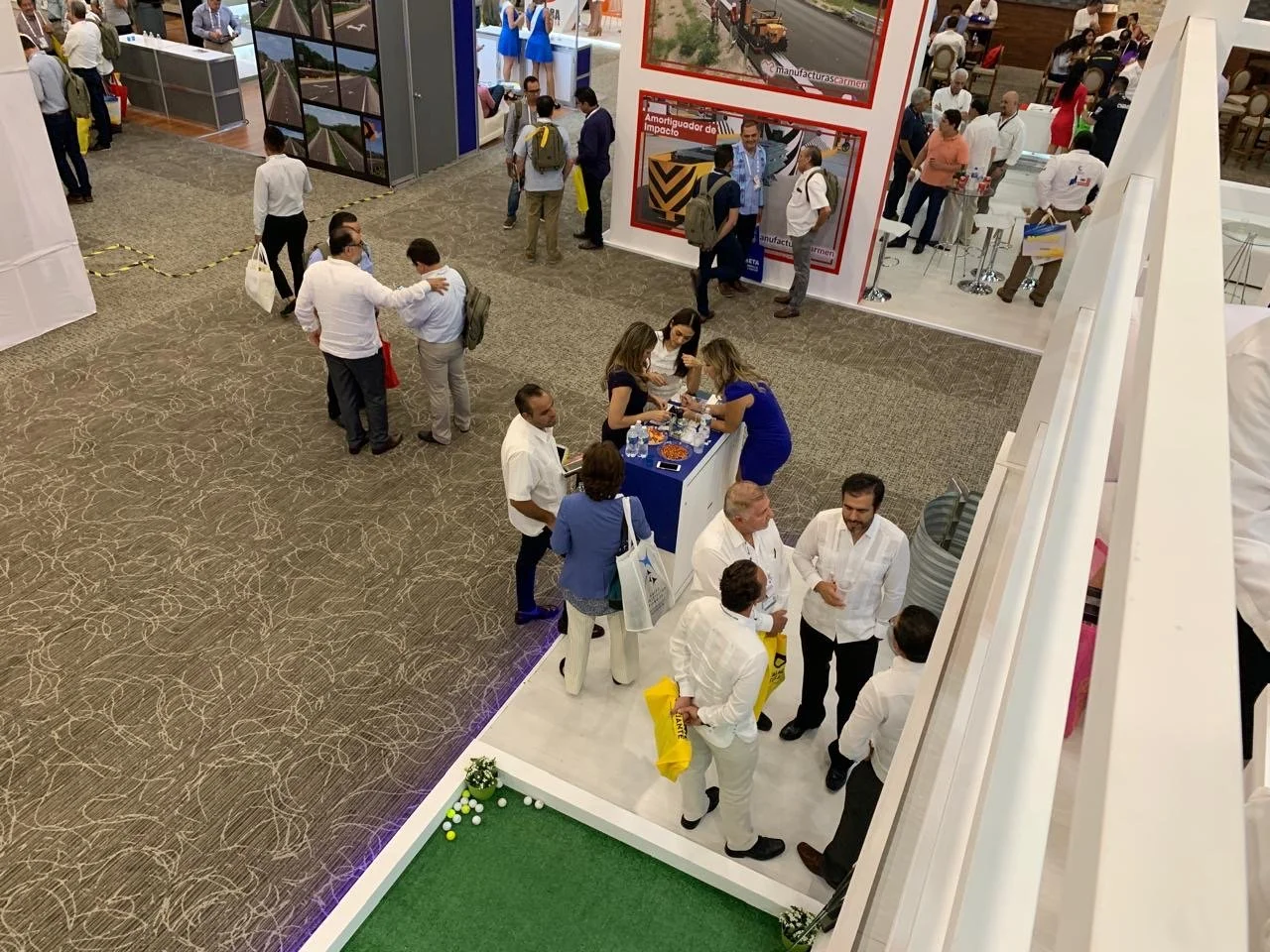 People attending a trade show or conference, engaging in conversations, and exploring booths. Some individuals are gathered around a blue table with snacks, while others walk around or stand in groups. The setting is an indoor exhibition hall with va