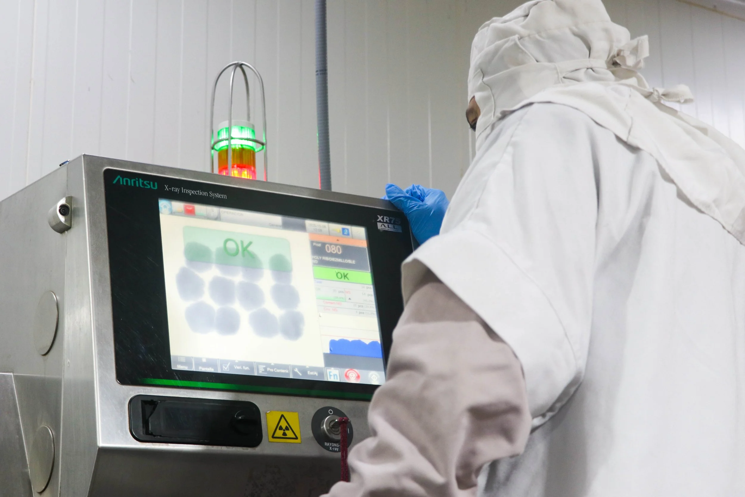A person dressed in protective clothing and gloves operating an X-ray inspection machine, with a screen displaying 'OK' and a green light indicating the status.