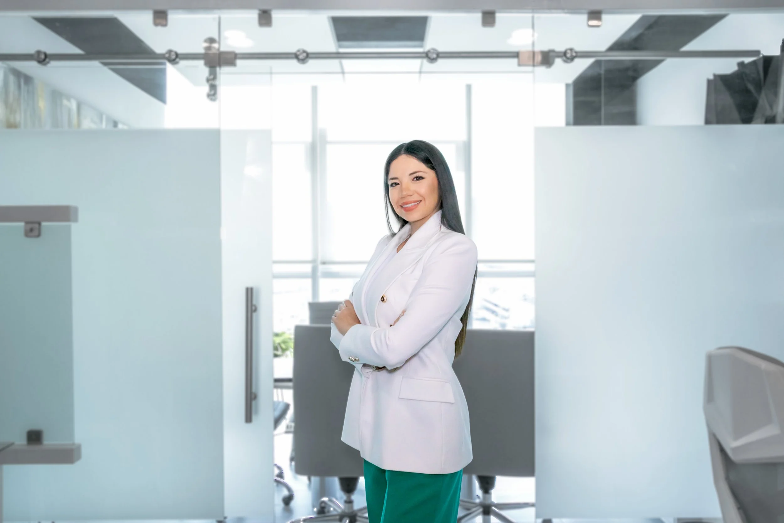 A woman in a white blazer and green pants standing with her arms crossed in a modern office space with large windows and high ceilings.