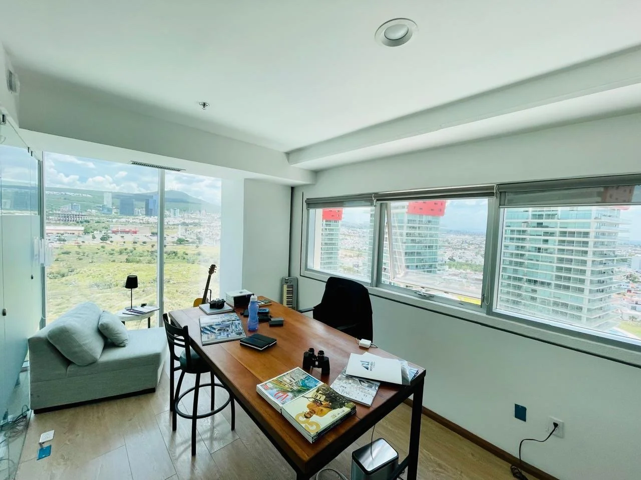 Modern office with large windows overlooking a city and hills, containing a wooden desk with books, electronics, and a water bottle, a black chair, a small sofa with pillows, and a guitar.
