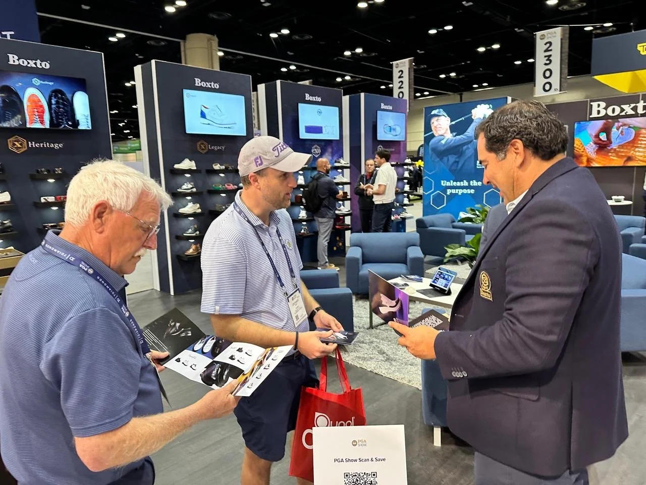 Three men are engaging in a conversation at a trade show booth for Bexto shoes, with displays of various shoes and digital screens in the background. One man is holding brochures, another is listening, and the third is reading a flyer, with seating a