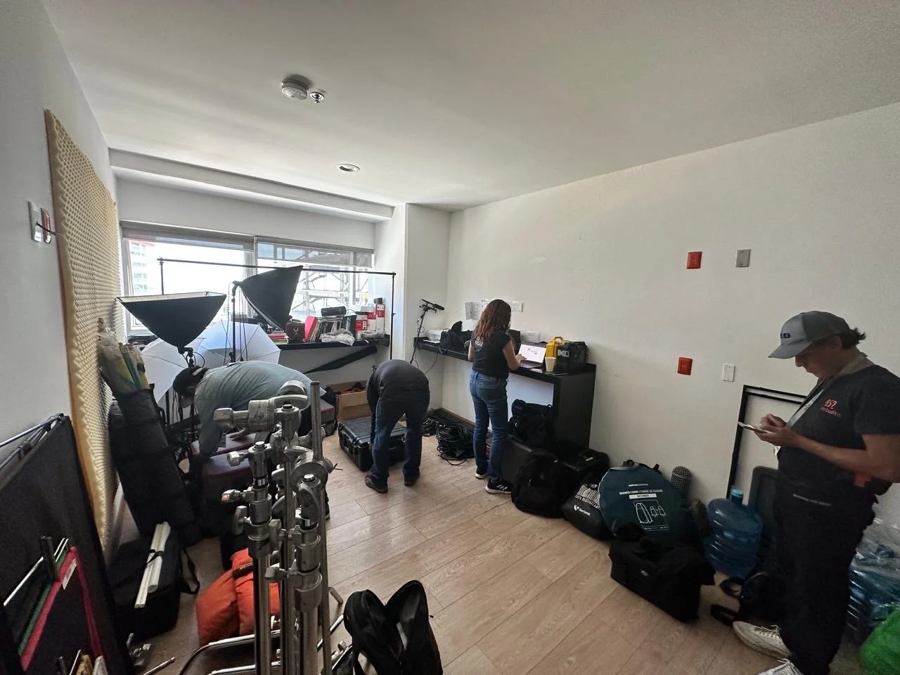 A small indoor room with three people preparing equipment, including lighting gear, bags, and water bottles, with a large window and technical supplies along the back wall.