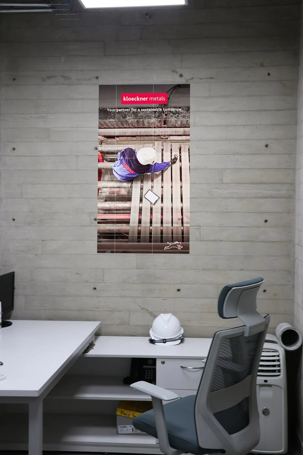 Office with a white desk, gray ergonomic chair, a white hard hat, and a poster on the wall featuring a worker inspecting metal pipes with the Kloeckner Metals logo.