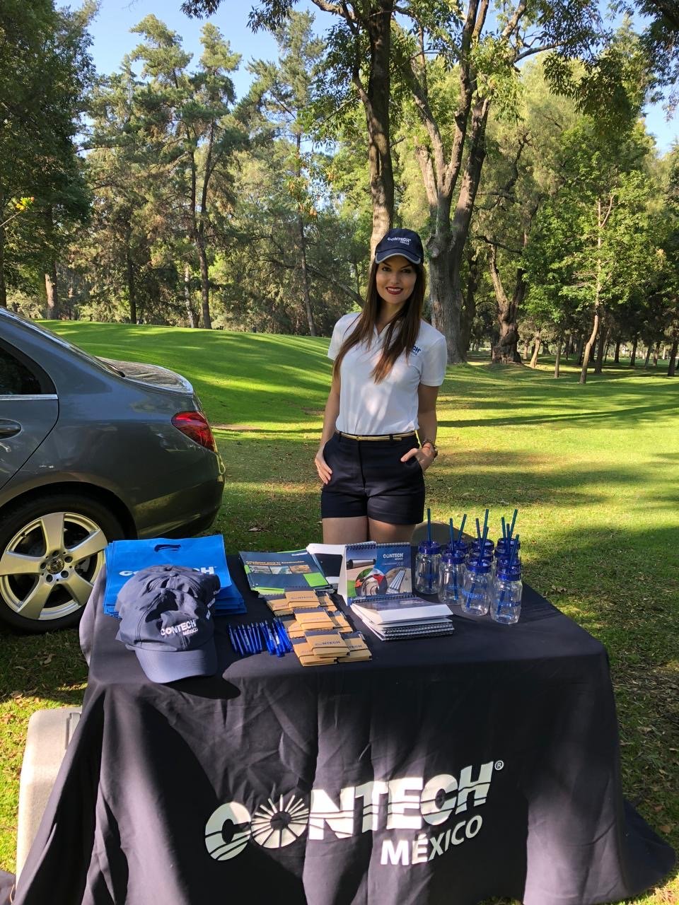 A woman stands at a promotional booth set up outdoors on a golf course, surrounded by trees, with a gray car partly visible on the left. The table is covered with a black cloth that has a white "CONTECH MEXICO" logo. Items on the table include caps, 
