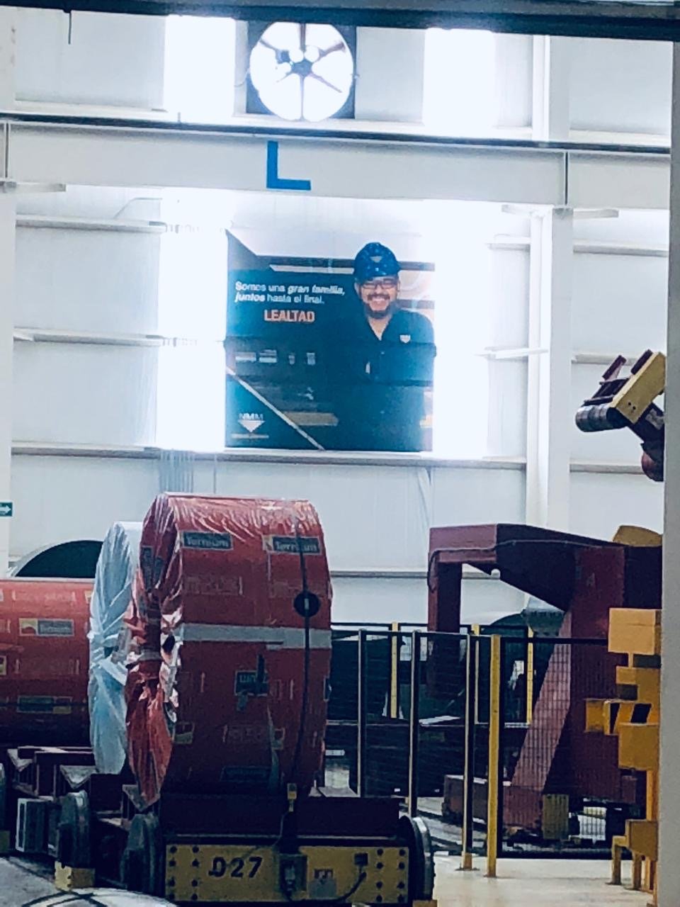 A store aisle with large red boxes or packages, some covered in plastic, and store equipment like yellow carts and black platforms. A large poster of a smiling man wearing a helmet and glasses is visible on the wall.