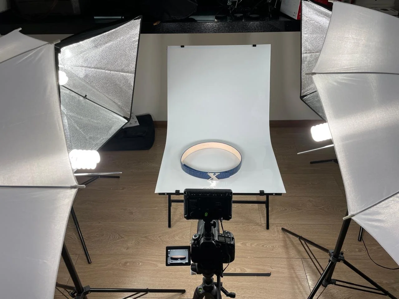 Photograph of a photography studio setup with a camera on a tripod aimed at a white background. The background features a belt with a metallic buckle placed on it, and three studio lights with reflective umbrellas illuminating the scene.