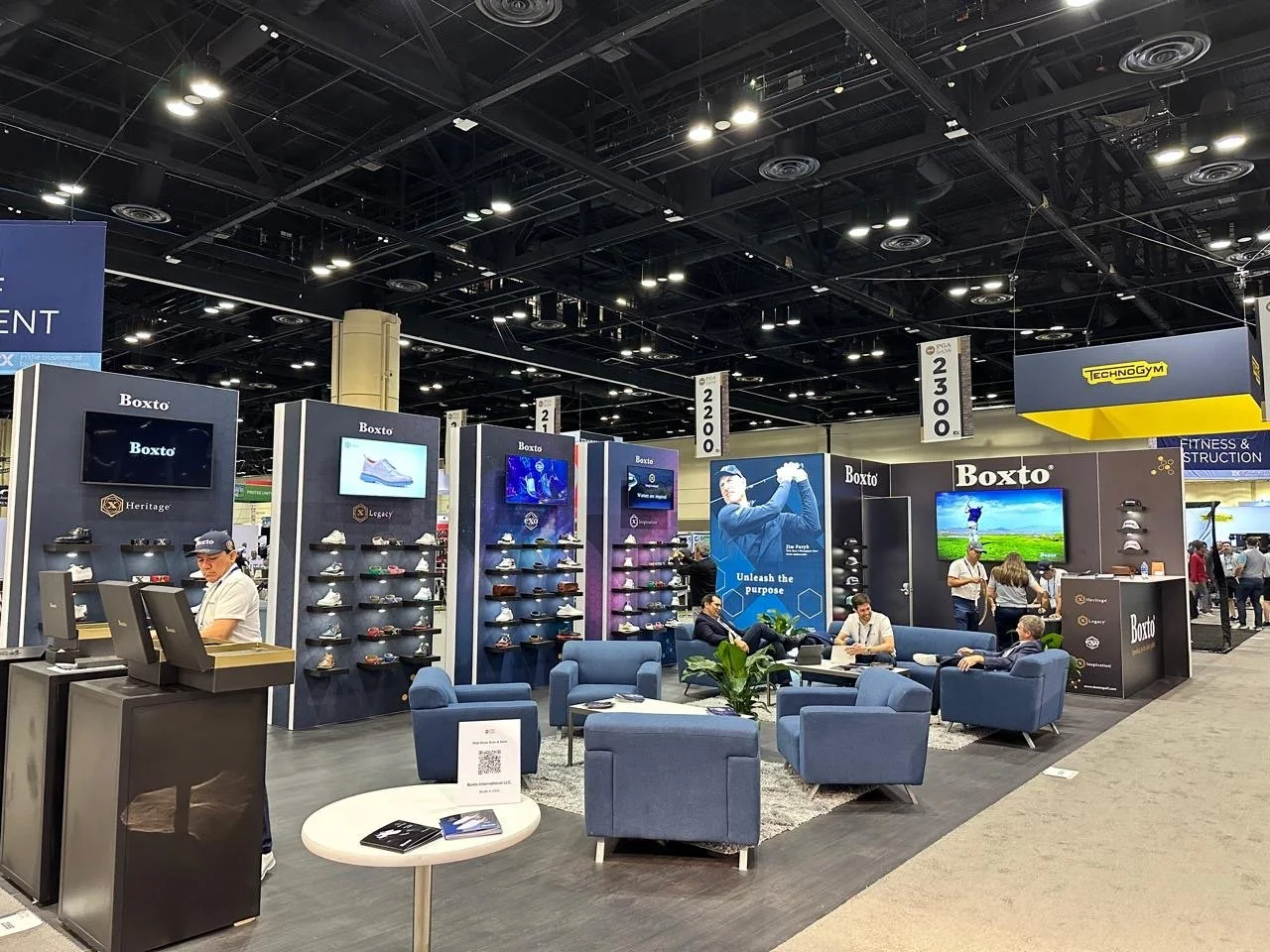Trade show booth displaying athletic shoes, seating area with people, large TV screens, promotional banners, and signage for Boxt and Technogym.