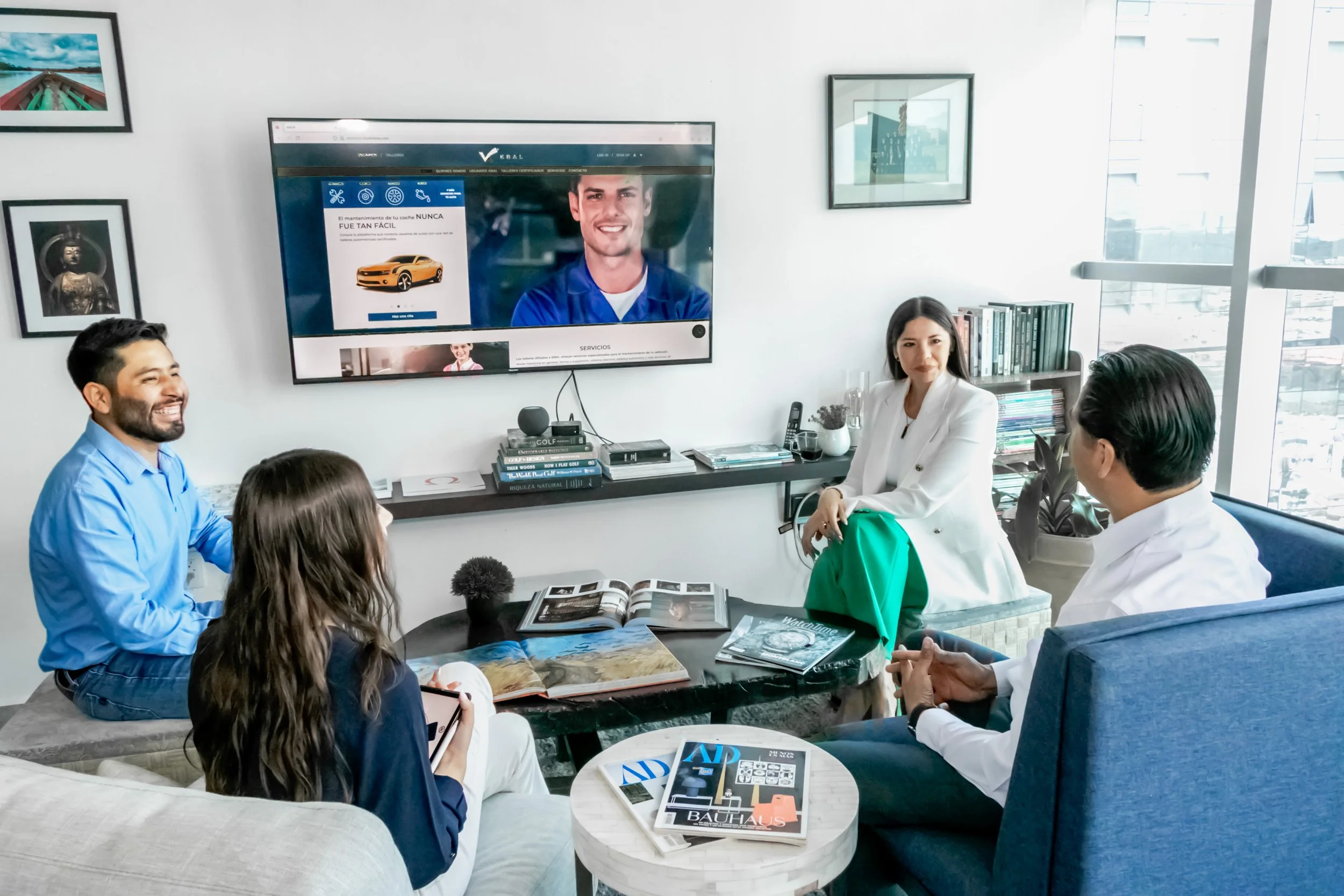 A group of five people in a modern office meeting room, sitting around coffee tables with magazines, in front of a large wall-mounted screen displaying a smiling man and a car website; they are engaged in a discussion.