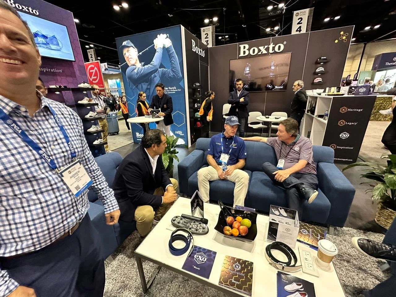 Trade show booth for Boxto featuring a large graphic of a golfer swinging a club, with several people engaging in discussions, sitting on a blue couch, and standing around in the booth area.