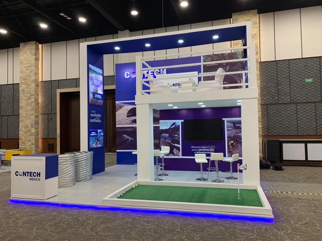 Trade show booth for CONTECH México with a two-tiered design, featuring seating, digital screens, and promotional banners in an indoor venue with decorative wall panels and carpeting.