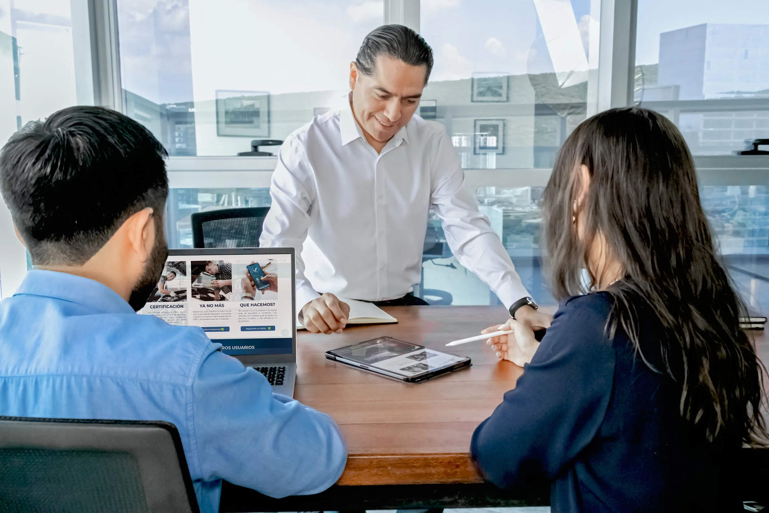 Business meeting in a modern office with three people, two sitting at a wooden table and one standing. The man standing is smiling and reviewing a tablet, while the man in a blue shirt and woman in a dark blouse are looking at a tablet on the table. 