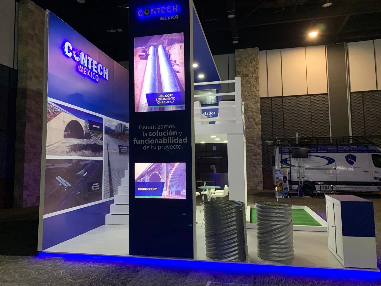 Exhibition booth for CONTECH Mexico featuring images of infrastructure projects and technology, including a large digital display, promotional signage, a small seating area, and a van in the background.