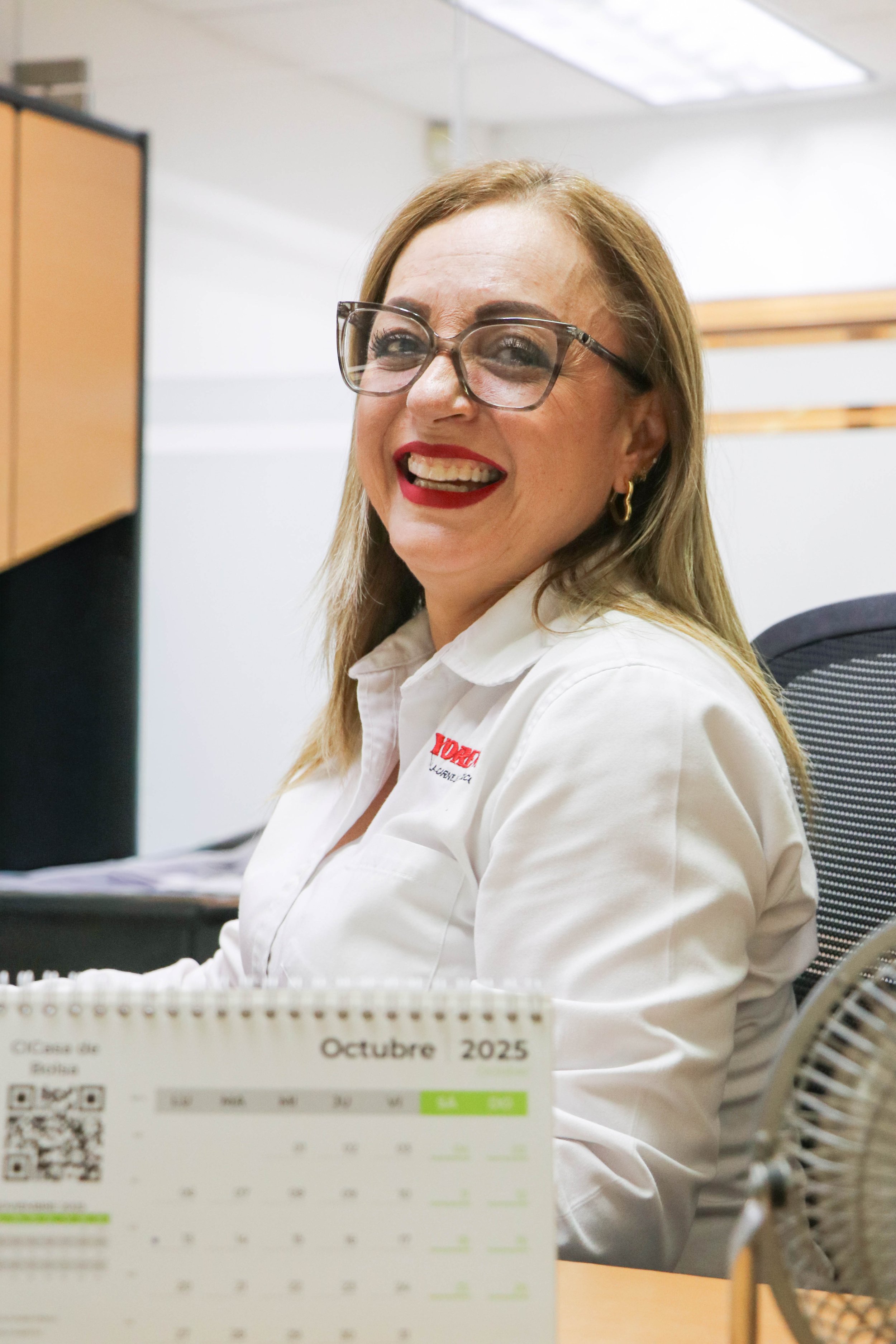 A woman with glasses and long blonde hair smiling at her desk in an office, wearing a white shirt with a logo, and a calendar on the desk showing October 2025.