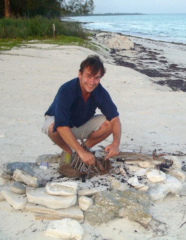 A man crouching on a sandy beach near the ocean, smiling at the camera, with a small fire pit made of rocks and driftwood in front of him.