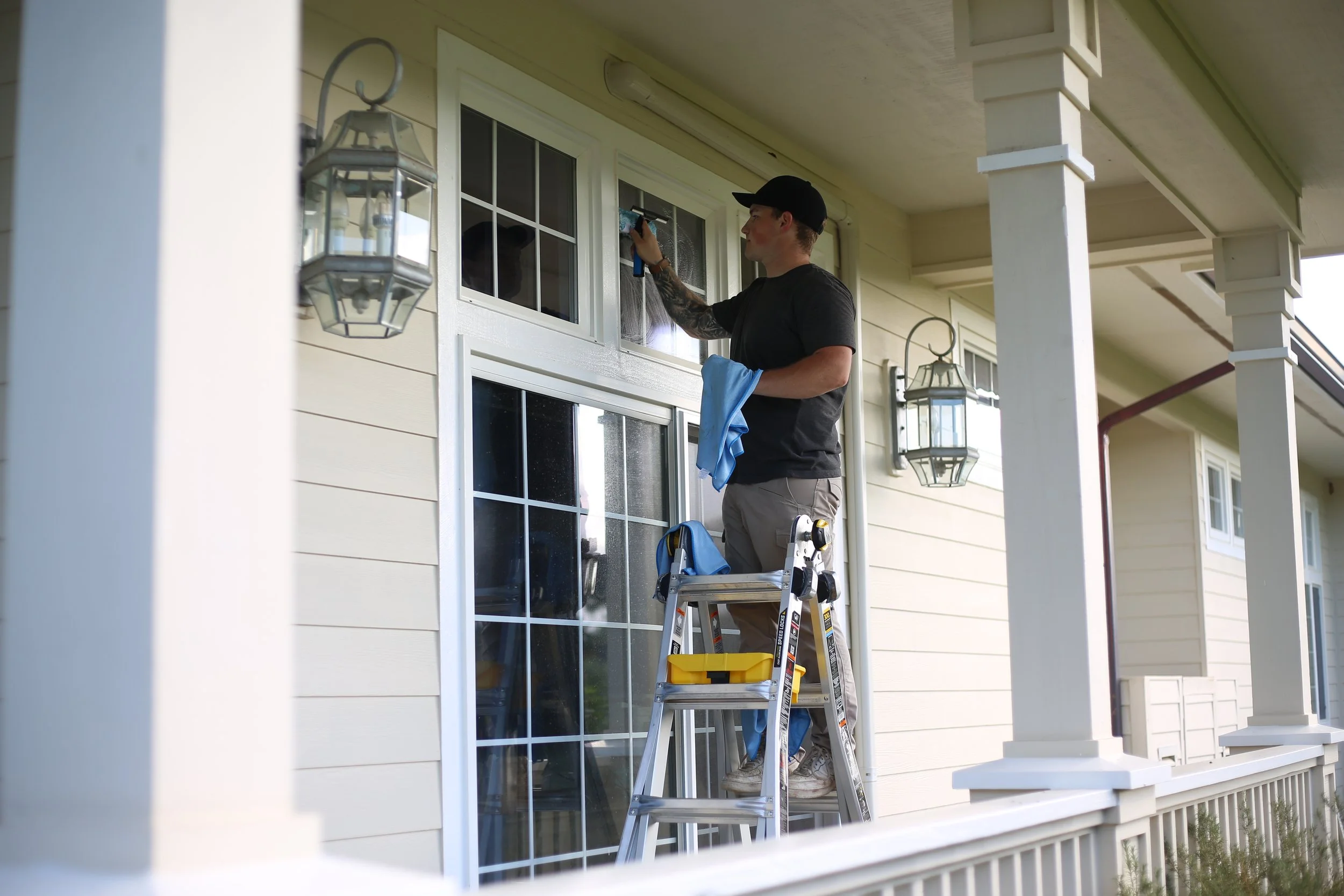 A man standing on a ladder cleaning a window on a house's exterior, holding a cloth in one hand and a spray bottle in the other.